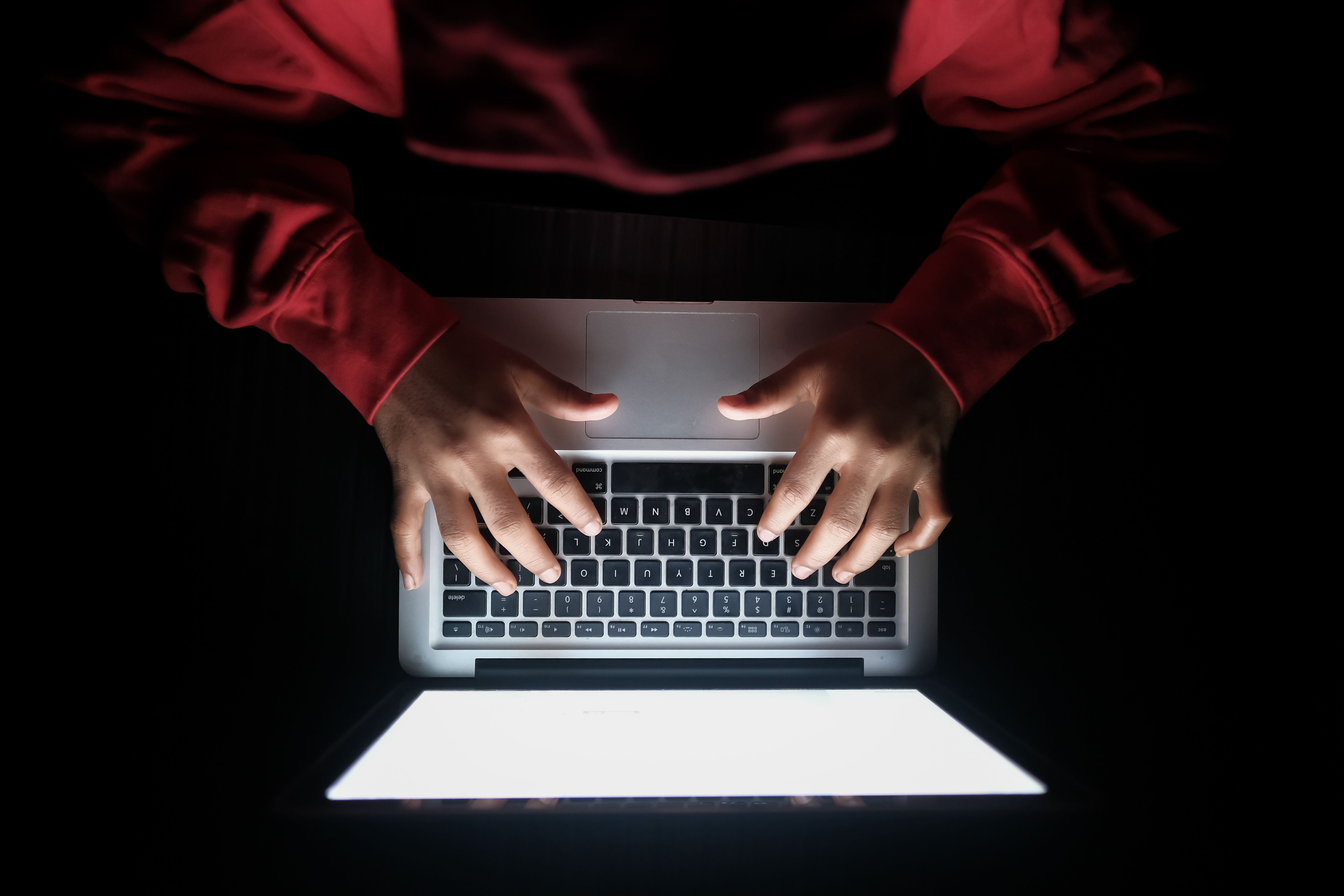 An overhead view of a person in a red hoodie typing on a laptop in a dark room.