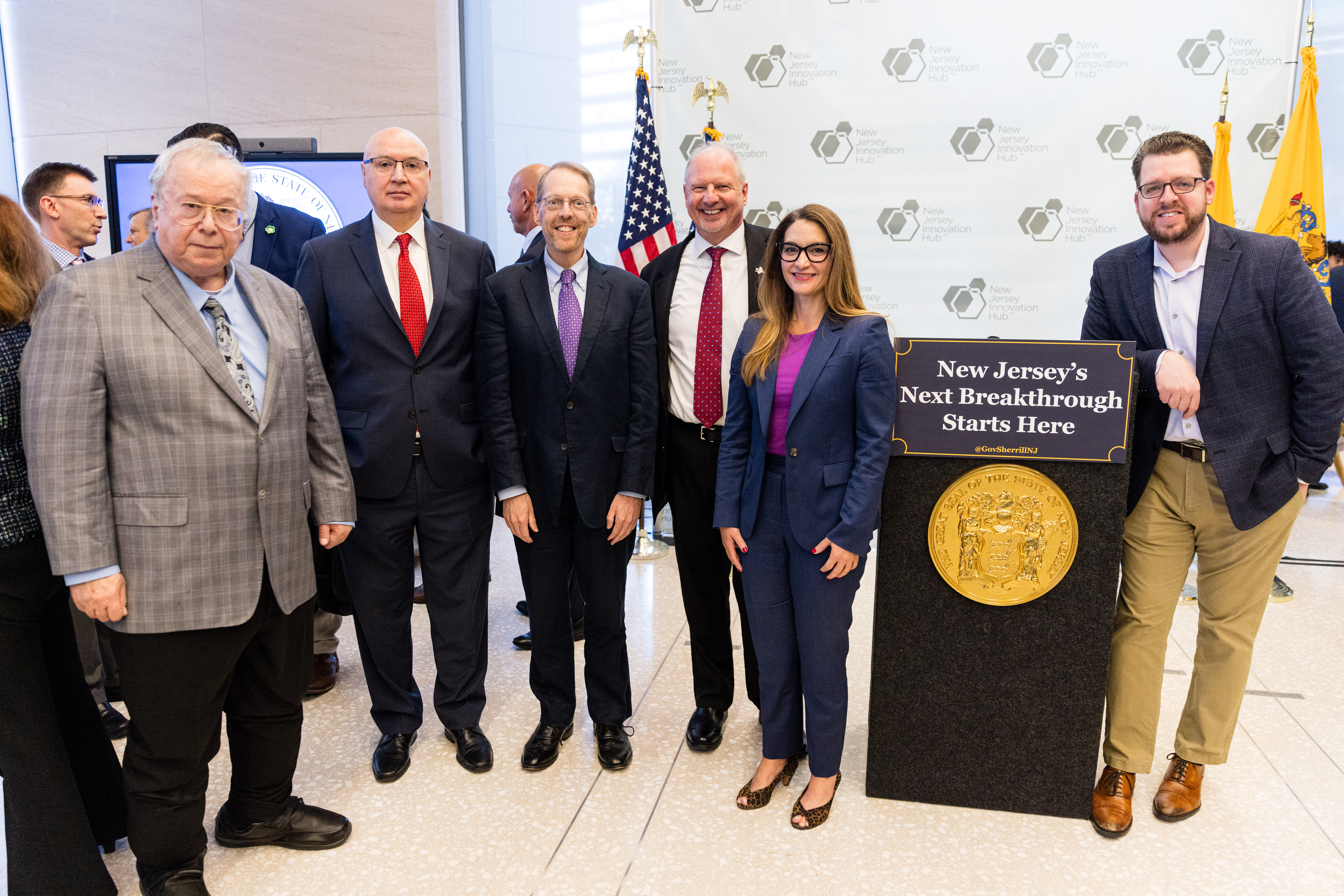 A group of five men and one woman stand around a lectern with a gold seal on the front and a sign reading "New Jersey's Next Breakthrough Starts Here"