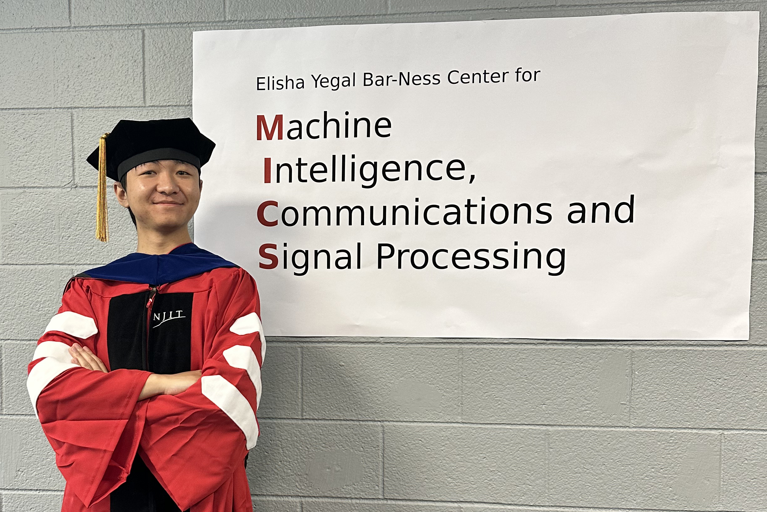 Yuan Zhao, wearing PhD regalia, stands next to a sign reading "Elisha Yegal Bar-Ness Center for Machine Intelligence, Communications and Signal Processing"