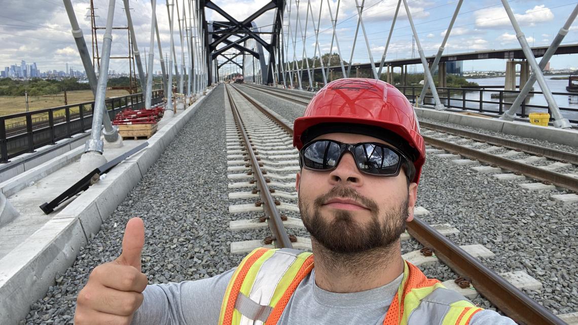 Enzo Suarez, wearing a hard hat and sunglasses, stands in front of railroad tracks giving a thumbs-up to the camera.