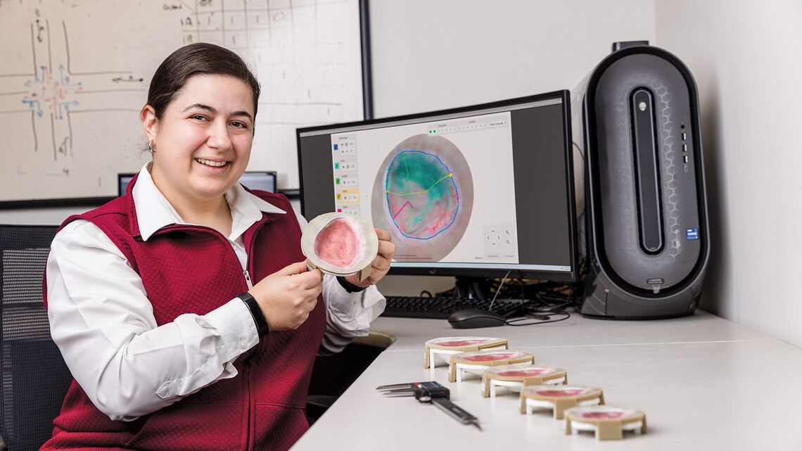 NJIT associate professor Salam Daher sits in front of a computer screen showing a digital wound model. She holds a 3D printed model of a skin anomaly.