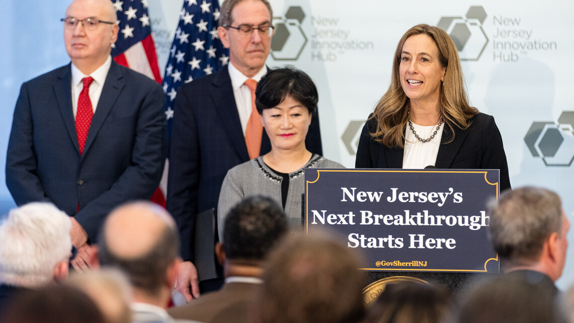 Governor Mikie Sherrill stands on a stage behind a podium, in front of a seated audience. A sign in front of the podium reads: "New Jersey's Next Breakthrough Starts Here"