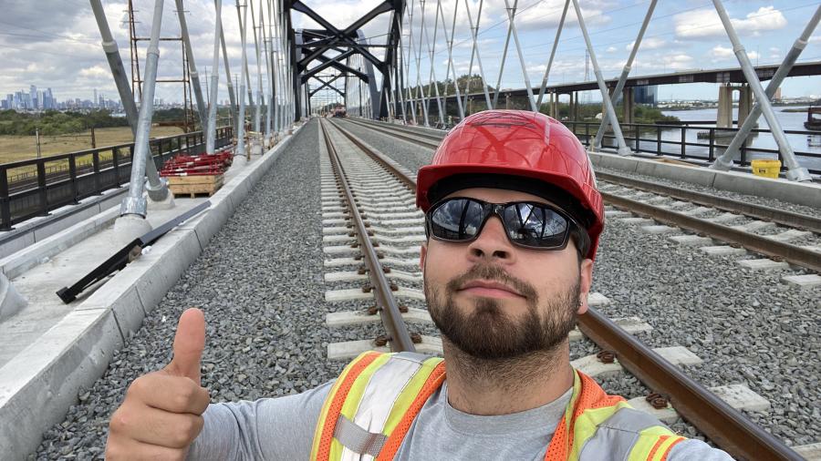 Enzo Suarez, wearing a hard hat and sunglasses, stands in front of railroad tracks giving a thumbs-up to the camera.