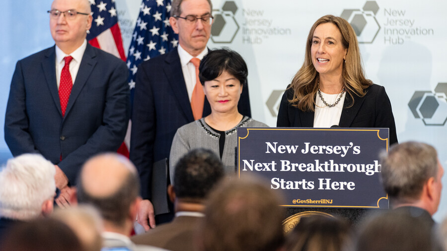 Governor Mikie Sherrill stands on a stage behind a podium, in front of a seated audience. A sign in front of the podium reads: "New Jersey's Next Breakthrough Starts Here"